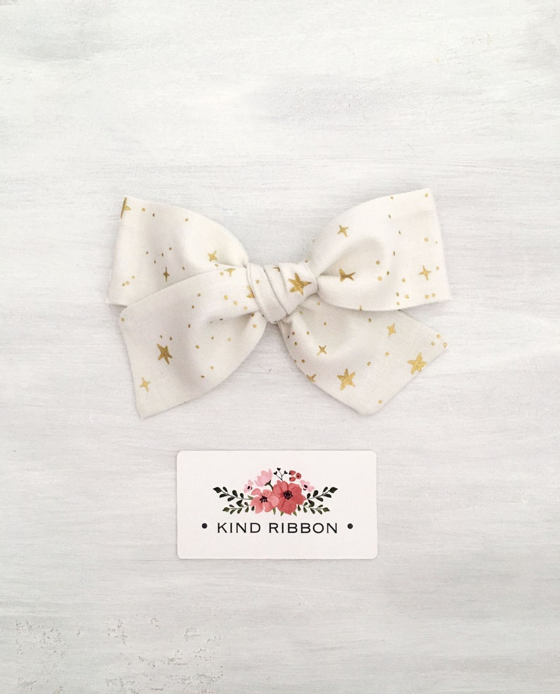 May include: A white fabric bow with gold stars and dots. The bow is tied in a knot and has a small tag with the text "KIND RIBBON" on it.
