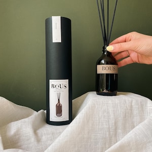 May include: A black cylindrical box with the word "BOUS" printed on it. The box contains a brown glass bottle with a black label that reads "BOUS Eco Diffuser with Essential Oils". The bottle is filled with a clear liquid and has black diffuser sticks inserted into it.