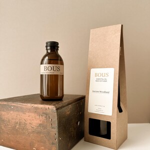 May include: A brown glass bottle of BOUS Ancient Woodland essential oil reed diffuser with a black cap, accompanied by a tall, light brown paper box featuring a white label with the text BOUS ESSENTIAL OIL REED DIFFUSER Ancient Woodland.