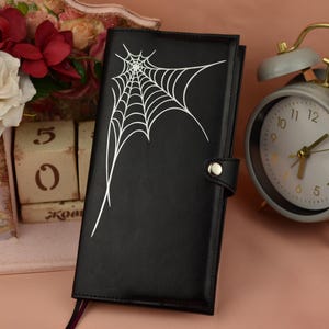 May include: A black rectangular notebook featuring a white spiderweb graphic on the front cover, secured with a metal snap closure. The notebook is positioned next to a gray analog alarm clock with gold accents and a decorative floral display.
