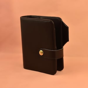 May include: A black leather wallet with a gold-colored button clasp. The wallet is open, revealing the interior. The background is a solid peach color.
