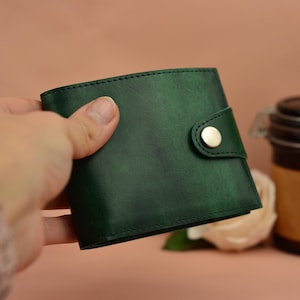May include: A dark green leather wallet with a snap closure. The wallet is held in a hand, with a jar and a flower in the background. The wallet has a textured appearance and a gold-colored snap.