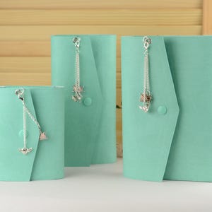May include: Three teal-colored journals of varying sizes. Each journal has a flap closure and a decorative silver chain with charms. The journals are arranged on a white surface with a wooden background.