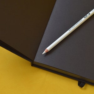 Pu&ograve; includere: Un taccuino nero con una matita bianca sopra. La matita ha la scritta "KOH-I-NOOR HARDTMUTH" stampata su di essa. Il taccuino &egrave; aperto e le pagine sono vuote.