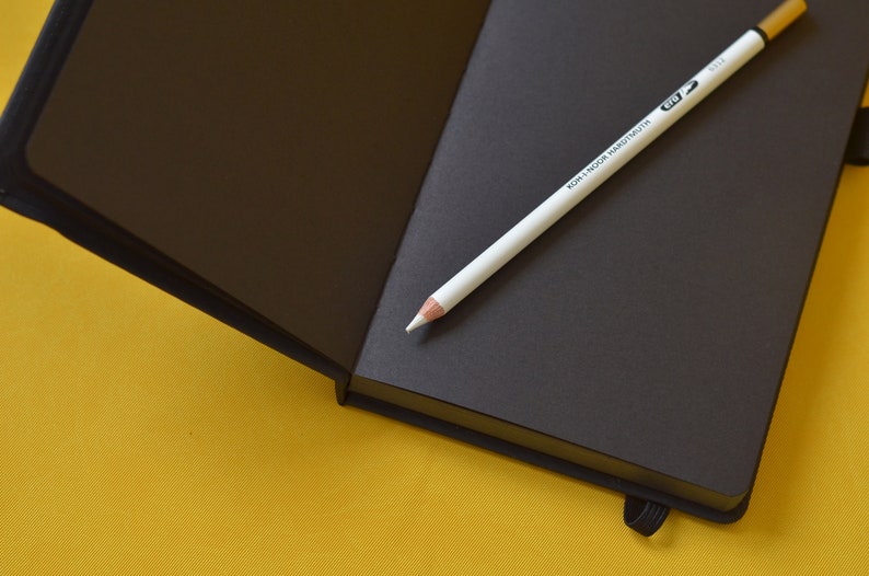 Pu&ograve; includere: Un taccuino nero con una matita bianca sopra. La matita ha la scritta "KOH-I-NOOR HARDTMUTH" stampata su di essa. Il taccuino &egrave; aperto e le pagine sono vuote.