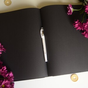 Puede incluir: Un cuaderno negro con un bol&iacute;grafo blanco abierto sobre una superficie blanca. Hay flores rosas a cada lado del cuaderno.