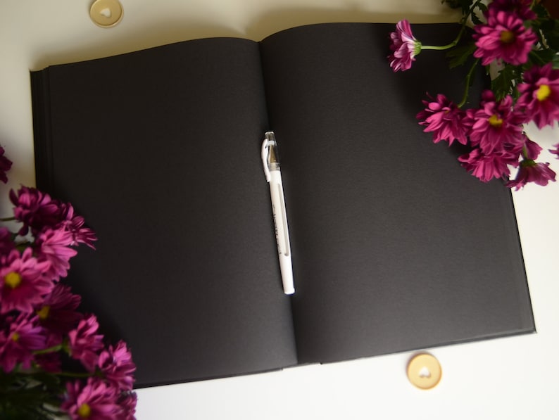 Puede incluir: Un cuaderno negro con un bol&iacute;grafo blanco abierto sobre una superficie blanca. Hay flores rosas a cada lado del cuaderno.