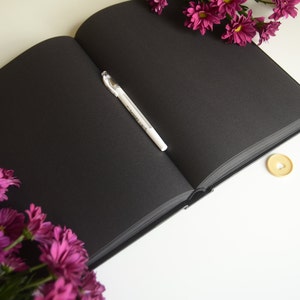 Puede incluir: Un cuaderno negro con un bol&iacute;grafo blanco abierto sobre una superficie blanca. El cuaderno est&aacute; rodeado de flores moradas.