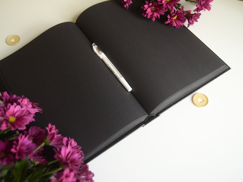 Puede incluir: Un cuaderno negro con un bol&iacute;grafo blanco abierto sobre una superficie blanca. El cuaderno est&aacute; rodeado de flores moradas.