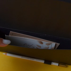 Pu&ograve; includere: Un taccuino in pelle nera con una tasca sul davanti. La tasca contiene diverse carte piegate con un motivo floreale. Il taccuino &egrave; aperto e l'interno &egrave; nero.