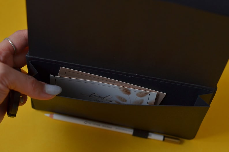 Pu&ograve; includere: Un taccuino in pelle nera con una tasca sul davanti. La tasca contiene diverse carte piegate con un motivo floreale. Il taccuino &egrave; aperto e l'interno &egrave; nero.