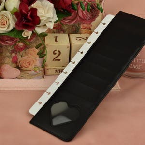May include: Black leather planner with a heart-shaped cutout, paired with a white, ring-bound section. The planner is set against a backdrop of flowers, a calendar, and a candle, creating a romantic aesthetic.