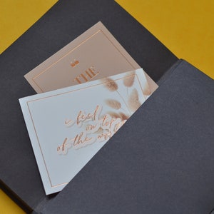 Pu&ograve; includere: Un taccuino nero con una tasca sul davanti. La tasca contiene due carte. Una carta &egrave; di colore marrone chiaro con la parola "THE" in lettere dorate. L'altra carta &egrave; bianca con un disegno floreale e il testo "I feel on top of the world" in lettere dorate.