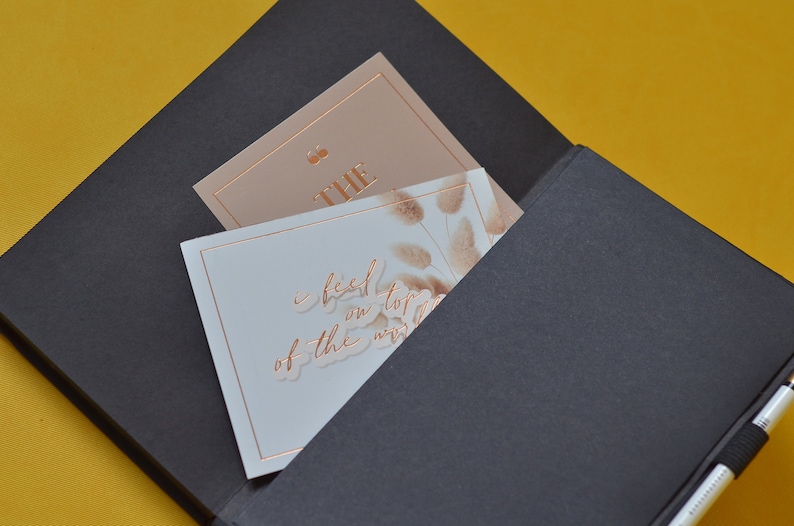 Pu&ograve; includere: Un taccuino nero con una tasca sul davanti. La tasca contiene due carte. Una carta &egrave; di colore marrone chiaro con la parola "THE" in lettere dorate. L'altra carta &egrave; bianca con un disegno floreale e il testo "I feel on top of the world" in lettere dorate.