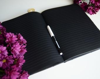 May include: A black lined journal with a white pen lying open on a white surface. A bouquet of purple flowers is in the lower left corner of the image.