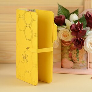 Pu&ograve; includere: Un portafoglio in pelle giallo con un motivo a nido d'ape e un design a forma di ape. Il portafoglio ha una chiusura a cinturino ed &egrave; aperto per rivelare l'interno.