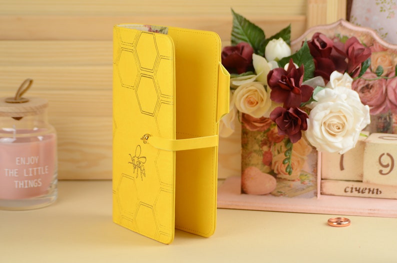 Pu&ograve; includere: Un portafoglio in pelle giallo con un motivo a nido d'ape e un design a forma di ape. Il portafoglio ha una chiusura a cinturino ed &egrave; aperto per rivelare l'interno.