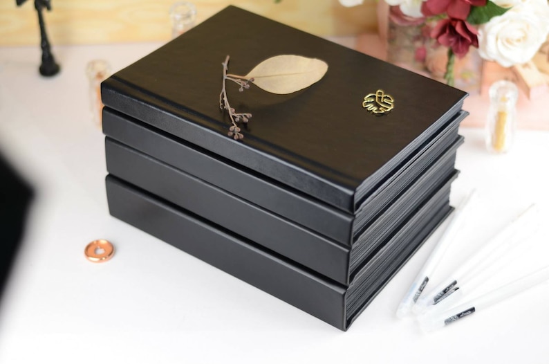 May include: A stack of four black hardcover journals with a gold leaf design on the top journal. The top journal has a dried leaf and a gold metal charm.