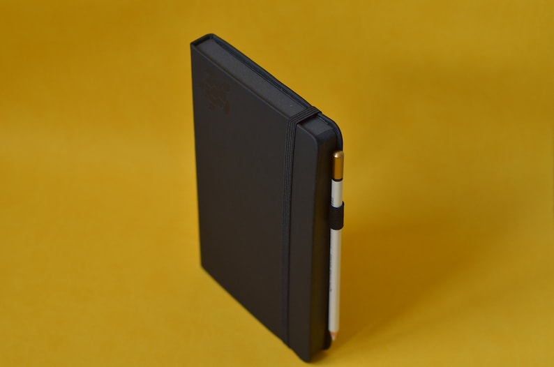A black faux leather notebook with an elastic closure and a white pencil tucked into the elastic band. Blank sketchbook, writing journal