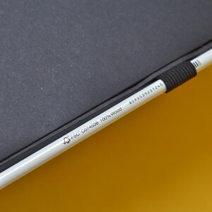 Pu&ograve; includere: Una matita bianca con una gomma nera e una punta dorata. La matita &egrave; etichettata con il testo "FSC C074598 100% Wood" e un numero "8595398231240". La matita &egrave; appoggiata su un taccuino nero.