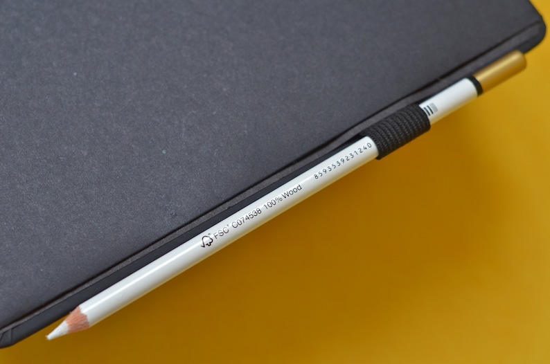 Pu&ograve; includere: Una matita bianca con una gomma nera e una punta dorata. La matita &egrave; etichettata con il testo "FSC C074598 100% Wood" e un numero "8595398231240". La matita &egrave; appoggiata su un taccuino nero.