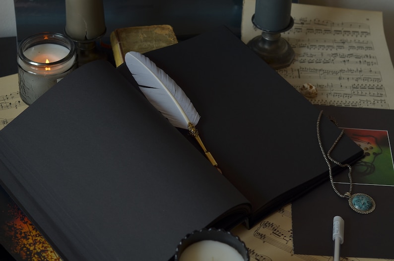 Puede incluir: Un diario encuadernado en cuero negro con una pluma de pluma blanca descansando sobre las p&aacute;ginas abiertas. El diario est&aacute; rodeado de velas, partituras musicales y un collar con un colgante de piedra turquesa.