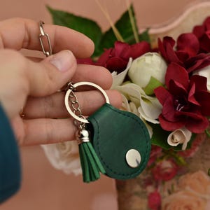 Pode incluir: Um porta-chaves de couro verde com corrente e borla prateadas. O porta-chaves tem um botão prateado redondo e é segurado em frente a um buquê de flores vermelhas e brancas.