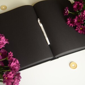 Puede incluir: Un diario de tapa dura negro con un bol&iacute;grafo blanco abierto sobre una superficie blanca. Hay flores rosas a la izquierda y a la derecha del diario. Hay dos peque&ntilde;as decoraciones doradas en forma de coraz&oacute;n en la superficie.