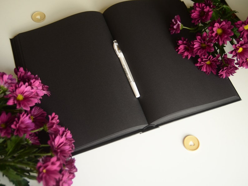 Puede incluir: Un diario de tapa dura negro con un bol&iacute;grafo blanco abierto sobre una superficie blanca. Hay flores rosas a la izquierda y a la derecha del diario. Hay dos peque&ntilde;as decoraciones doradas en forma de coraz&oacute;n en la superficie.