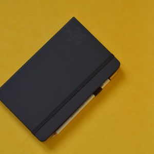 Pu&ograve; includere: Un taccuino nero con una chiusura elastica nera e una matita che sporge dalla parte superiore. Il taccuino &egrave; su una superficie gialla.