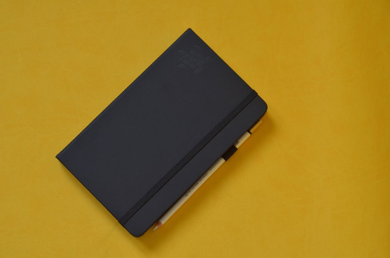 Pu&ograve; includere: Un taccuino nero con una chiusura elastica nera e una matita che sporge dalla parte superiore. Il taccuino &egrave; su una superficie gialla.