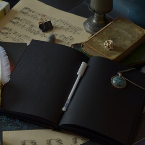 May include: A black leather-bound journal with a white pen lying open on a table. The journal is surrounded by candles, a feather quill, sheet music, and a necklace with a turquoise pendant.