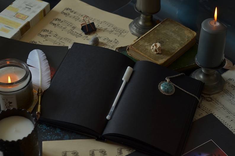 May include: A black leather-bound journal with a white pen lying open on a table. The journal is surrounded by candles, a feather quill, sheet music, and a necklace with a turquoise pendant.
