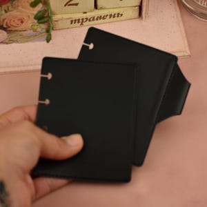 May include: A pair of black, square-shaped leather notebooks with three ring binder holes. The notebooks are held in a hand, with a watch visible on the wrist. The background is a soft pink color.