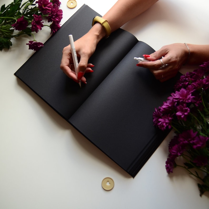 Puede incluir: Un cuaderno negro con un bol&iacute;grafo blanco y una botella transparente de l&iacute;quido sobre una superficie blanca con flores moradas.