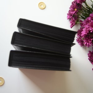 May include: A stack of four black hardcover books on a white surface. The books are arranged in a staggered pattern, with the top book slightly offset from the bottom book. There are three small, round, yellow buttons scattered around the books.
