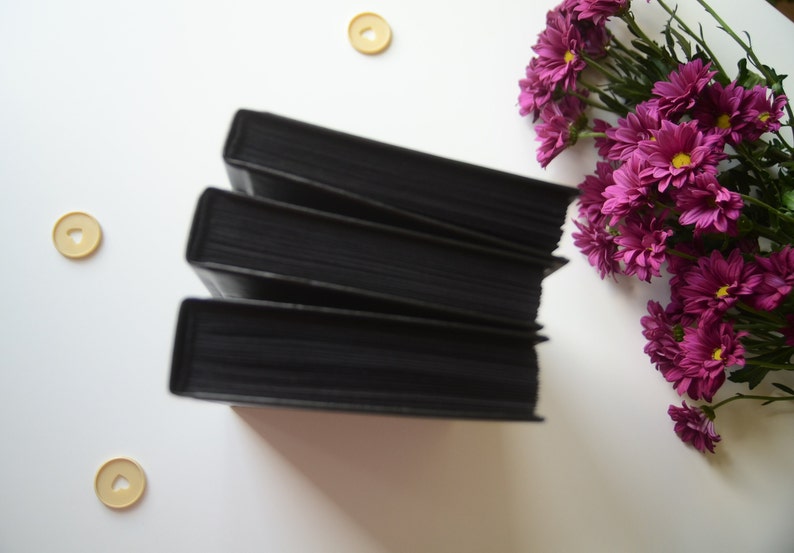 May include: A stack of four black hardcover books on a white surface. The books are arranged in a staggered pattern, with the top book slightly offset from the bottom book. There are three small, round, yellow buttons scattered around the books.