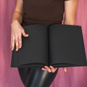 Puede incluir: Una persona sosteniendo un libro negro cerrado frente a un fondo rosa.