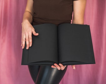 Può includere: Una persona tiene in mano un libro nero chiuso davanti a uno sfondo rosa.