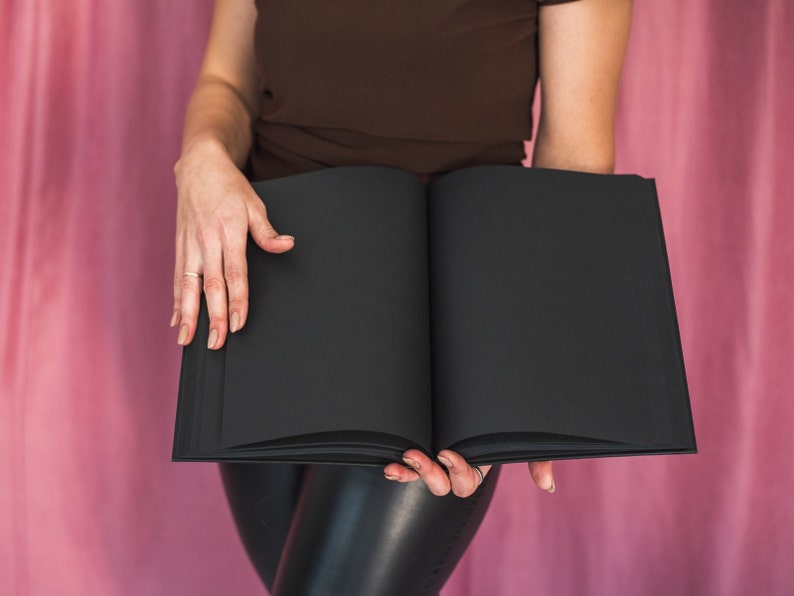 Puede incluir: Una persona sosteniendo un libro negro cerrado frente a un fondo rosa.