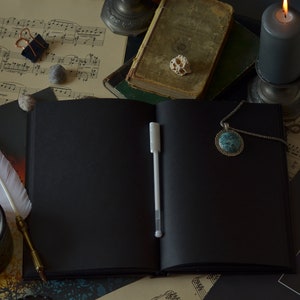 May include: A black leather bound journal with a white pen, a white feather quill, and a lit candle on a table with sheet music and a book.