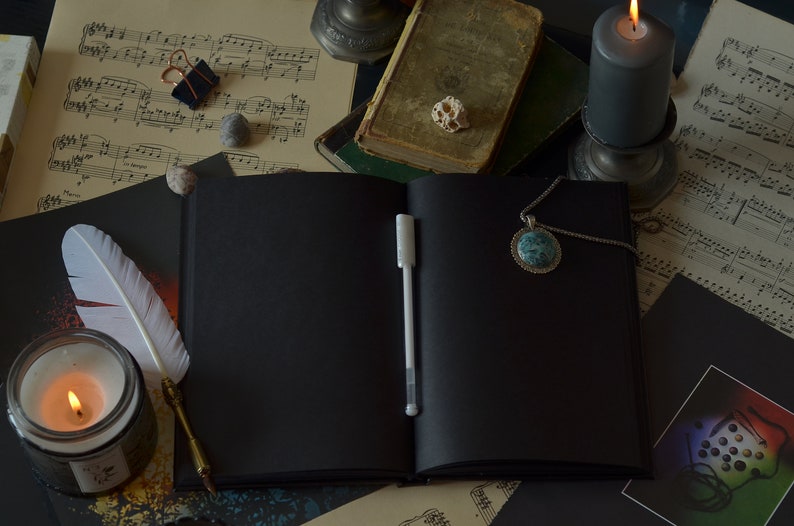 May include: A black leather bound journal with a white pen, a white feather quill, and a lit candle on a table with sheet music and a book.