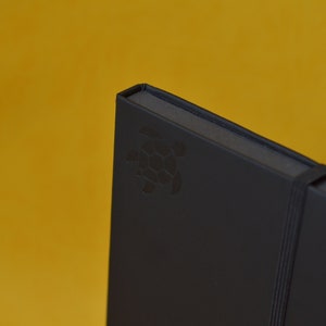 Pu&ograve; includere: Un taccuino nero con un logo di tartaruga in rilievo sulla copertina. Il taccuino ha una chiusura con elastico nero e una penna dorata attaccata alla copertina.