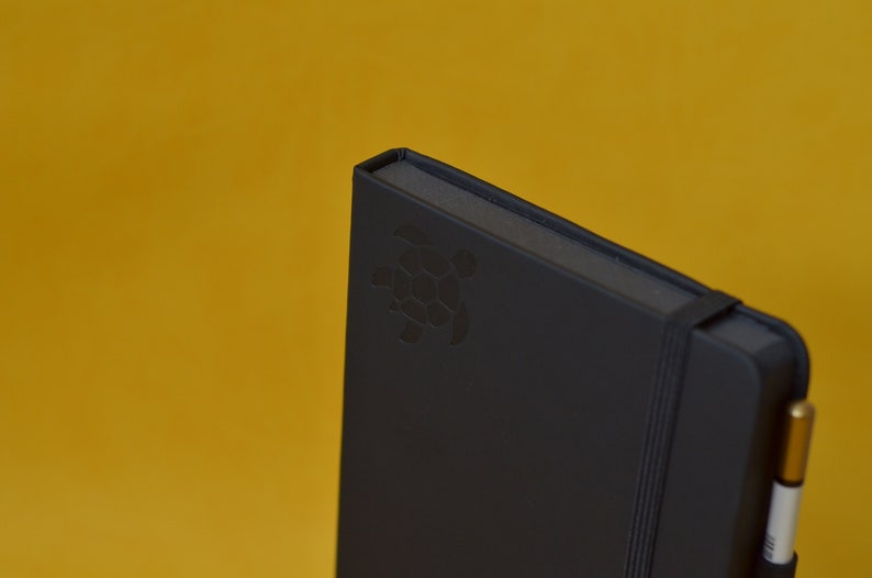 Pu&ograve; includere: Un taccuino nero con un logo di tartaruga in rilievo sulla copertina. Il taccuino ha una chiusura con elastico nero e una penna dorata attaccata alla copertina.