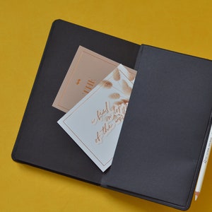 Pu&ograve; includere: Un taccuino nero con una penna dorata e due carte all'interno. La carta superiore &egrave; beige con la parola "the" scritta in oro. La carta inferiore &egrave; bianca con una scritta dorata che dice "I feel that I've been lost in the world".