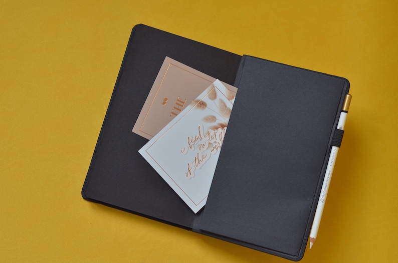 Pu&ograve; includere: Un taccuino nero con una penna dorata e due carte all'interno. La carta superiore &egrave; beige con la parola "the" scritta in oro. La carta inferiore &egrave; bianca con una scritta dorata che dice "I feel that I've been lost in the world".