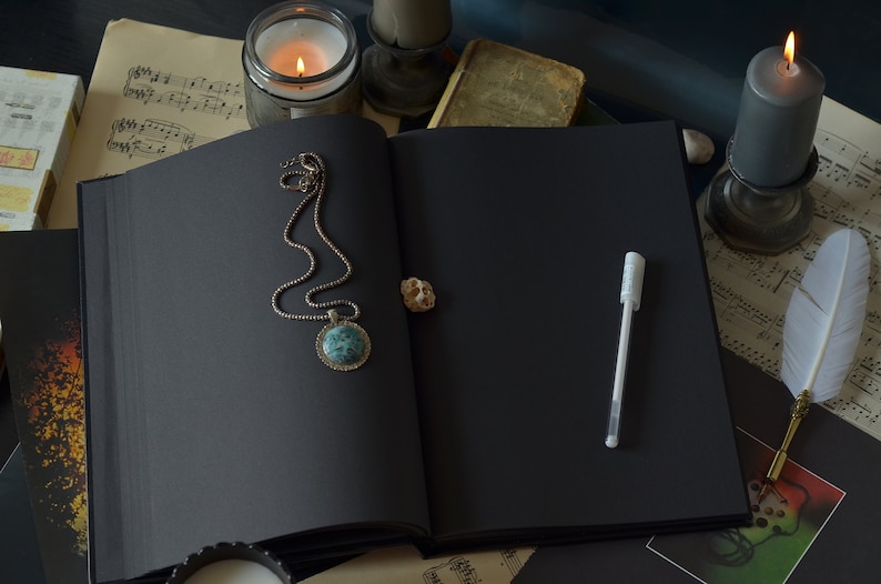 Puede incluir: Un diario negro encuadernado en cuero con un collar de cadena plateada con un colgante turquesa, un bol&iacute;grafo blanco y un bol&iacute;grafo de pluma blanca sobre una mesa con partituras y velas.