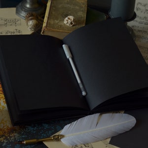 May include: An open black journal with a white pen lying on the open pages. A white feather quill pen is lying on a sheet of music with a silver necklace with a turquoise stone pendant.