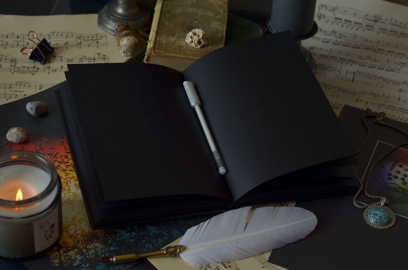 May include: An open black journal with a white pen lying on the open pages. A white feather quill pen is lying on a sheet of music with a silver necklace with a turquoise stone pendant.