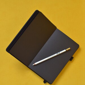 Pu&ograve; includere: Un taccuino nero con una matita bianca aperta su una superficie gialla.